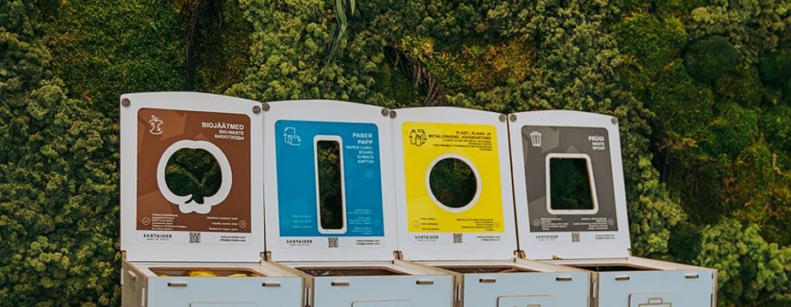 Sustainable waste sorting station with color-coded compartments for paper, packaging, bio, and mixed waste, placed against a green moss wall.